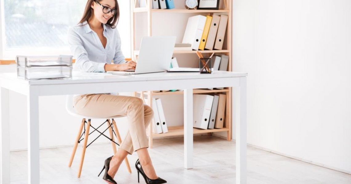 Photo of businesswoman dressed in white shirt and wearing glasses sitting in her office and using laptop. Looking aside.